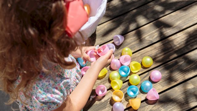 A child with a pink bow picks up colorful plastic Easter eggs scattered on a wooden deck, wearing a floral dress and short sleeves.