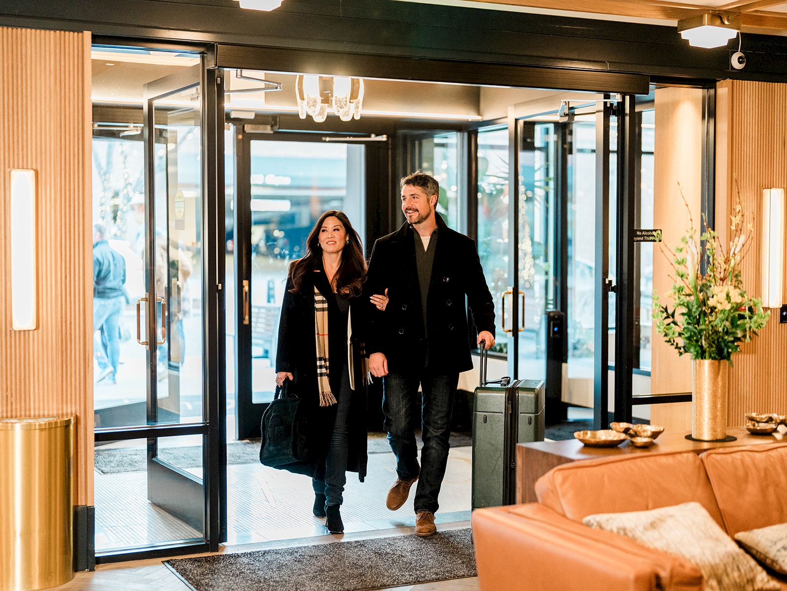 A stylish couple enters a modern lobby, carrying a suitcase, with warm lighting, glass doors, and a comfy seating area nearby.