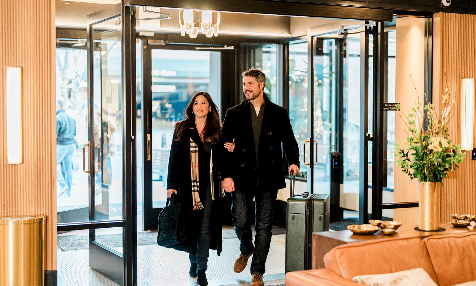 A stylish couple enters a modern lobby, carrying a suitcase, with warm lighting, glass doors, and a comfy seating area nearby.