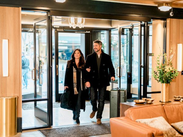 A stylish couple walks through a hotel lobby entrance, carrying coats and bags, bright modern decor with warm tones and plants.