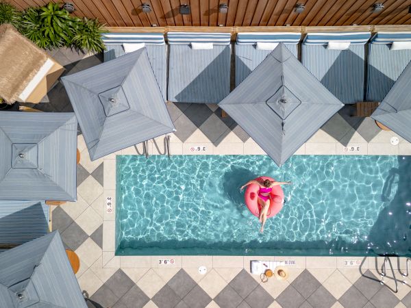 A rectangular rooftop pool with blue water, surrounded by gray umbrellas and a deck, featuring a person on a pink float in the center.