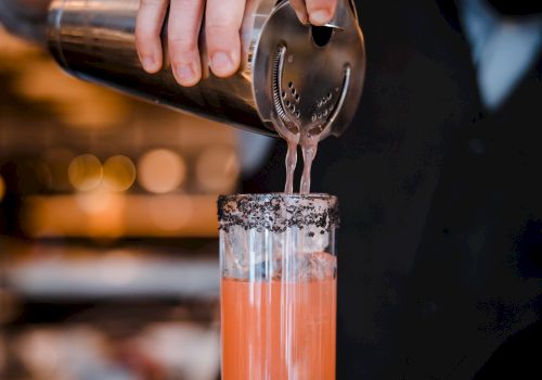 A bartender pours a pink cocktail through a strainer, creating a frothy, colorful drink in a tall glass with a bar backdrop.