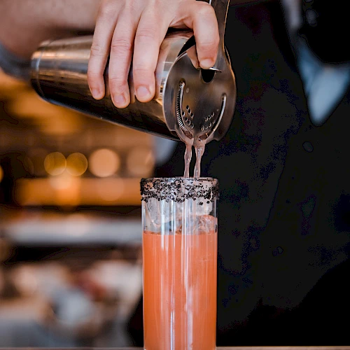A bartender pours a pink cocktail through a strainer, creating a frothy, colorful drink in a tall glass with a bar backdrop.