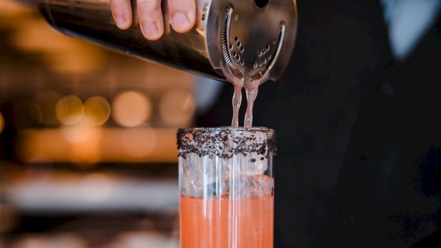 A bartender pours a pink cocktail through a strainer, creating a frothy, colorful drink in a tall glass with a bar backdrop.