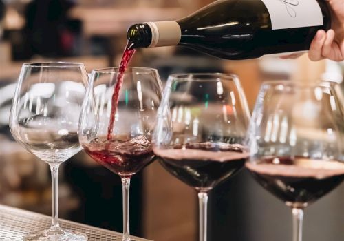 Pouring red wine into glasses at a bar, with a blurred background and a hand tilting the bottle to color the first glass.