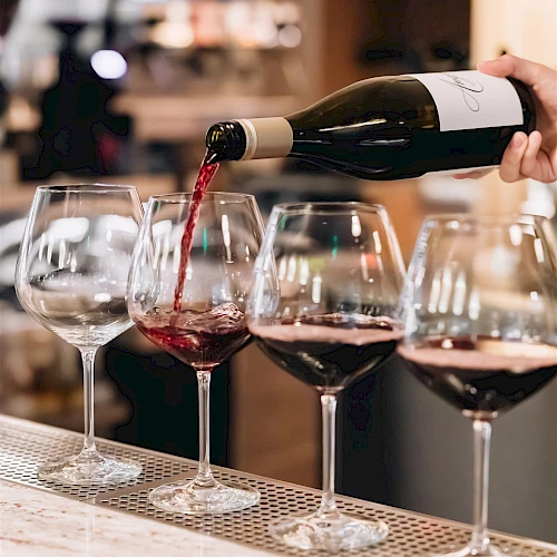 Pouring red wine into glasses at a bar, with a blurred background and a hand tilting the bottle to color the first glass.