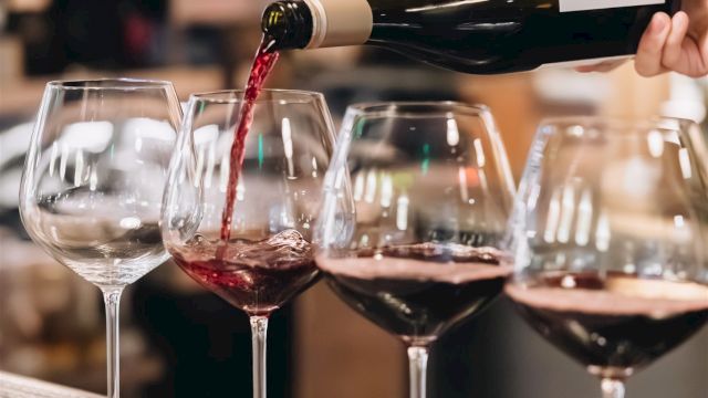 Pouring red wine into glasses at a bar, with a blurred background and a hand tilting the bottle to color the first glass.