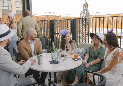 A group of friends in stylish outfits chat and laugh around a white round table on a sunny rooftop patio, drinks in hand, enjoying a moment.