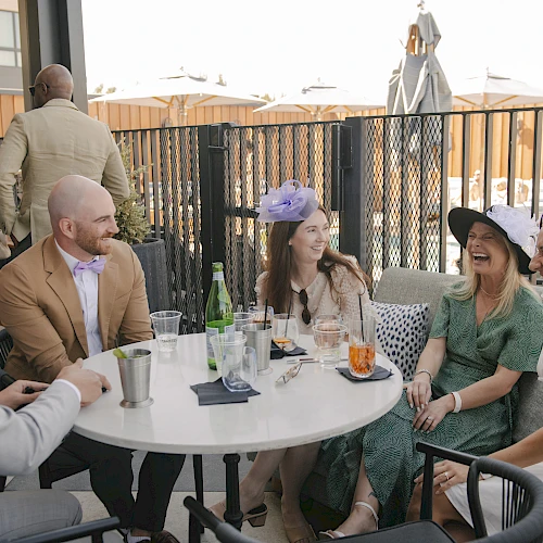 A group of friends in stylish outfits chat and laugh around a white round table on a sunny rooftop patio, drinks in hand, enjoying a moment.