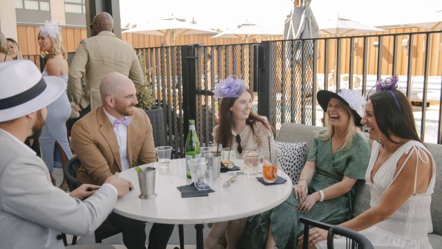 A group of friends in stylish outfits chat and laugh around a white round table on a sunny rooftop patio, drinks in hand, enjoying a moment.