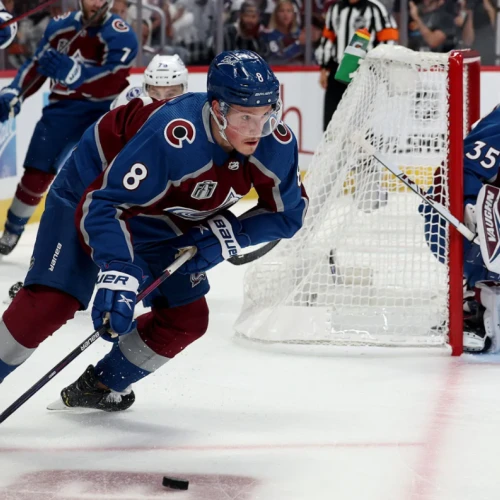Two hockey players chase the puck near the goal, with a goalie guarding the net as spectators watch intently.