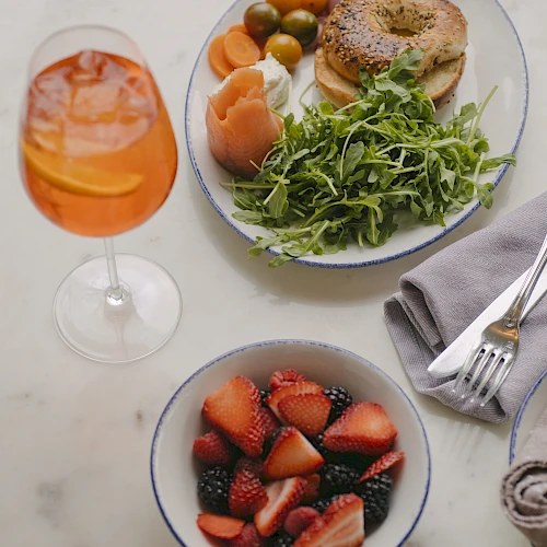 A brunch setup with a glass of orange drink, a plate of bagel, greens, olives, and fruit, plus a bowl of mixed berries on a marble table.