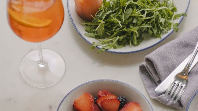 A brunch setup with a glass of orange drink, a plate of bagel, greens, olives, and fruit, plus a bowl of mixed berries on a marble table.