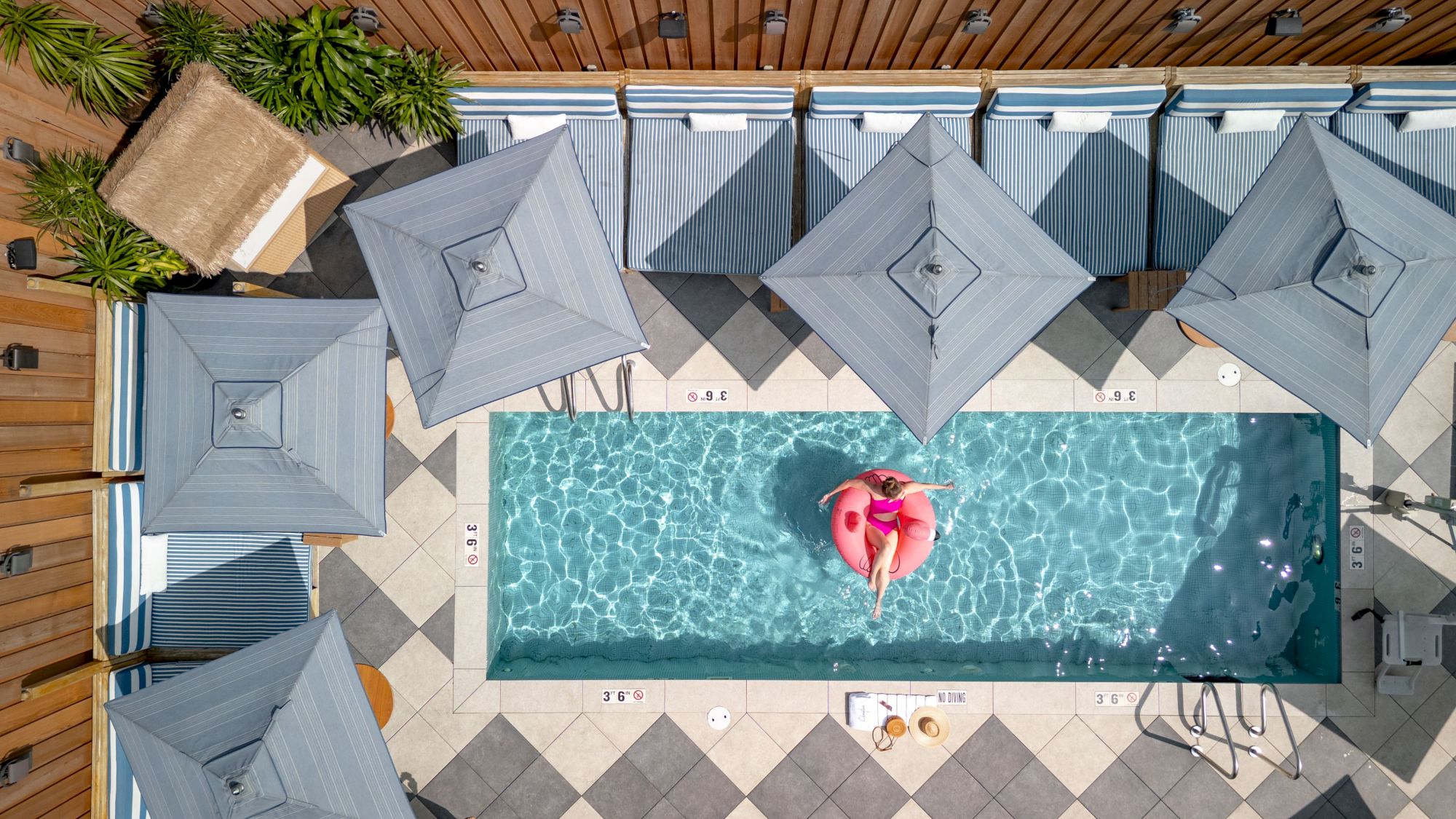 Aerial view of a rectangular pool with clear blue water, surrounded by gray umbrellas and lounge chairs on a tiled deck. A person lounges on a pink float.