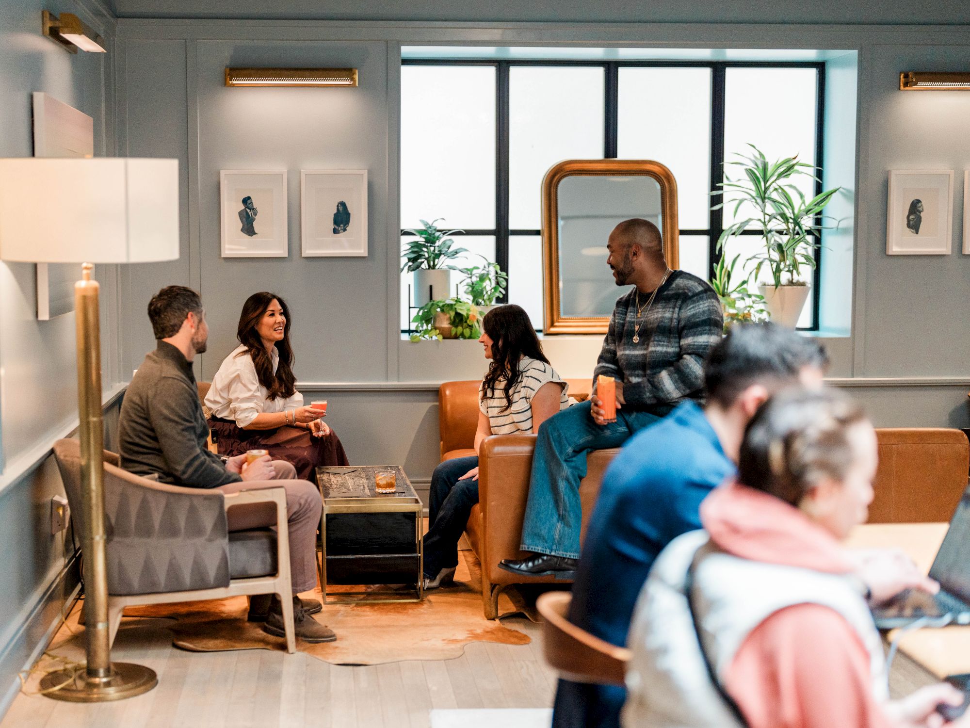A cozy lounge with people chatting and working on laptops, sunlight from a large window, plants, and modern decor.