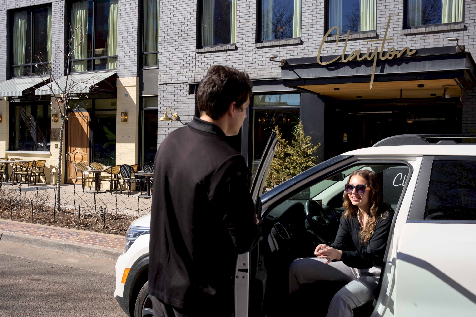 A man and woman are in a car conversation on a street with cafe seating and a storefront in the background.