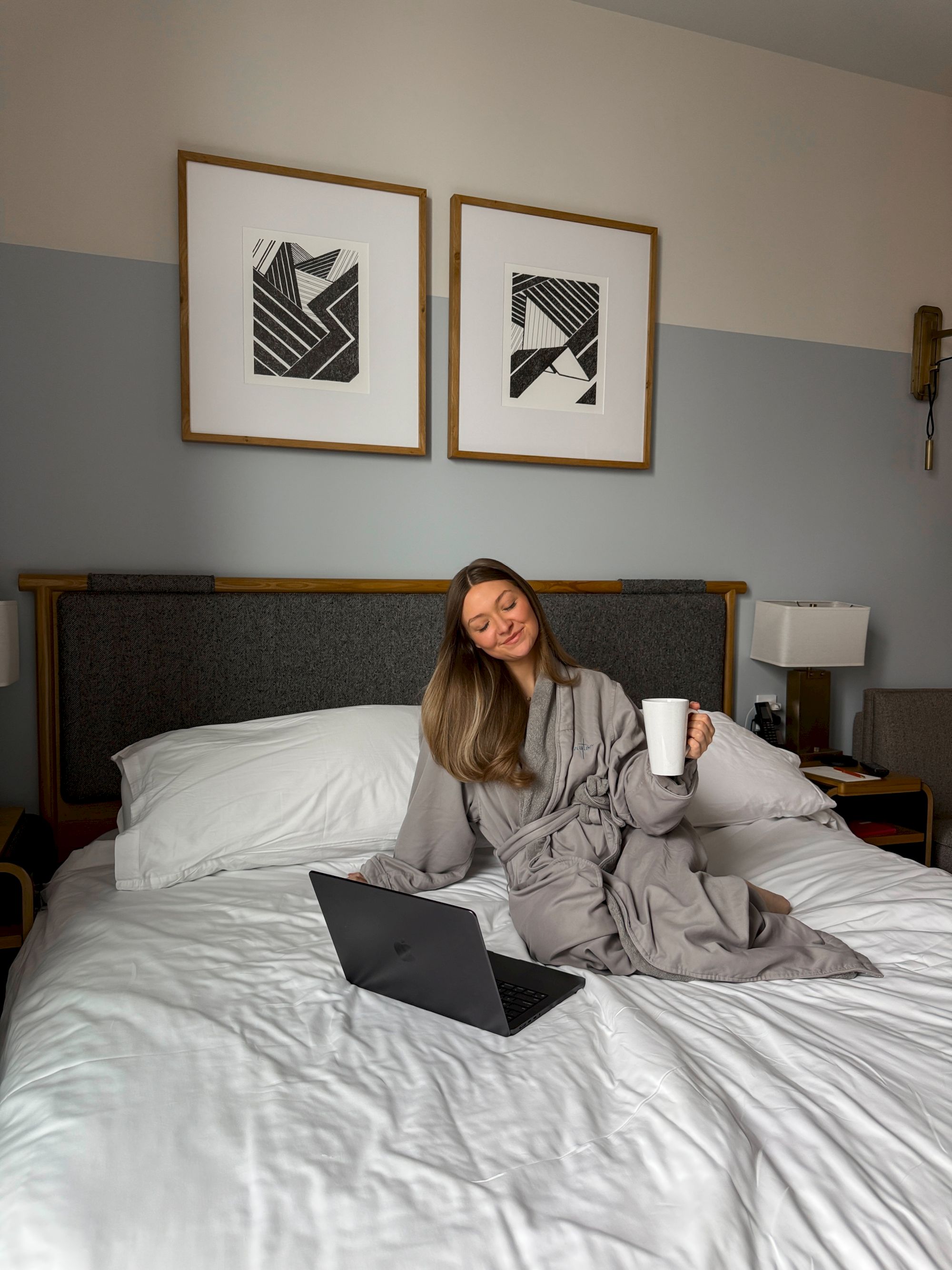 A woman sits on a bed with a laptop, wearing a gray robe, holding a cup, in a cozy bedroom with framed wall art and soft lighting.