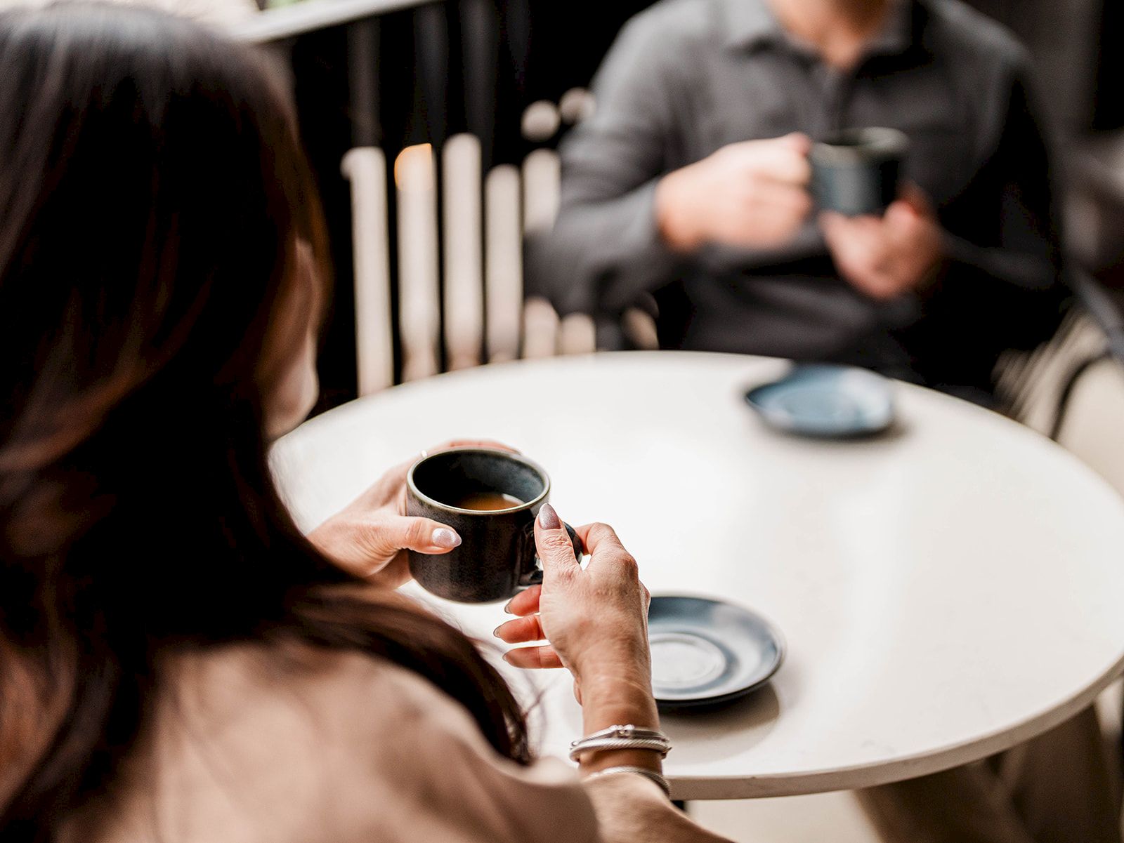 Two people sit at a cafe table; a woman holds a small black cup while a man across smiles and enjoys a drink.