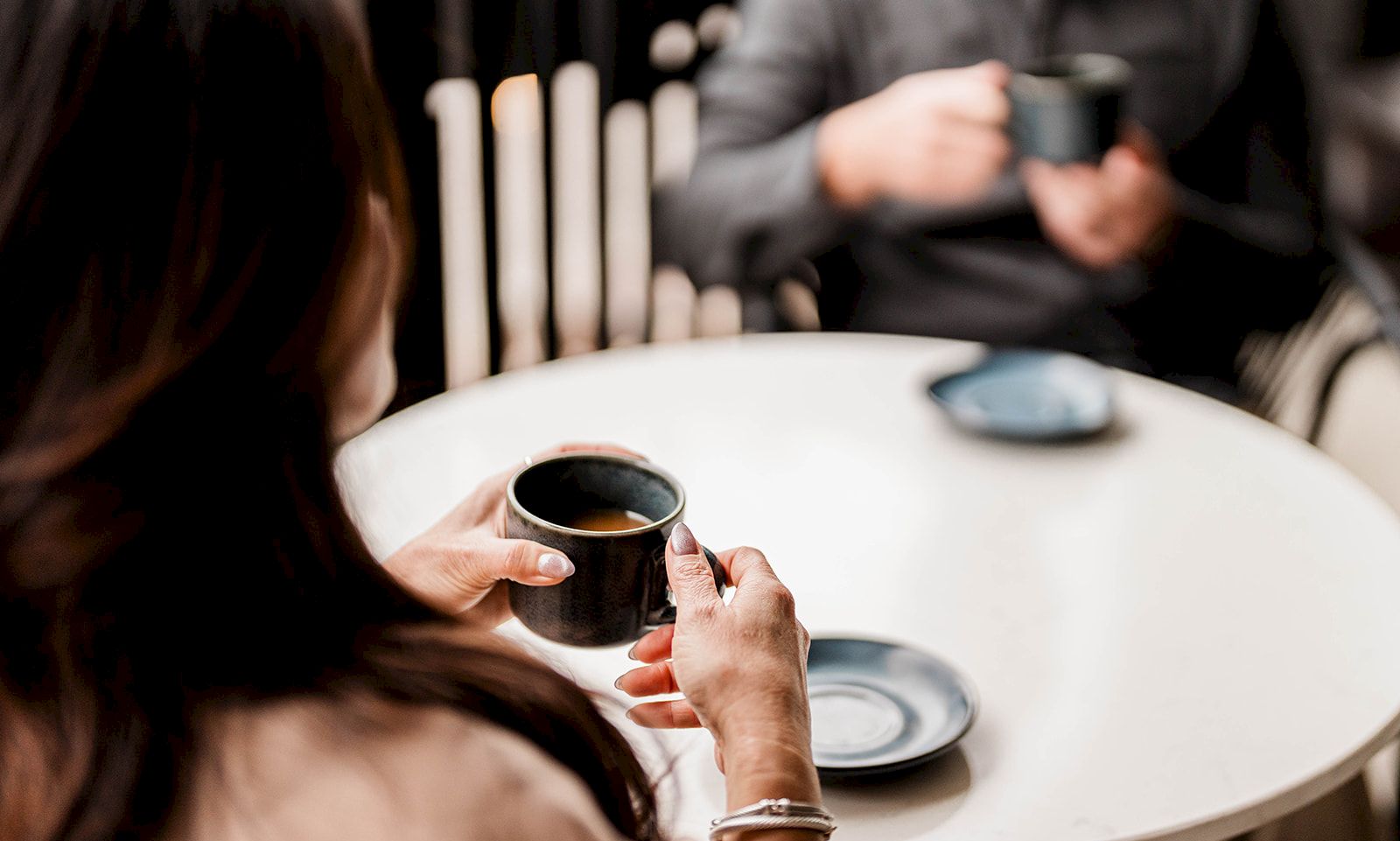 Two people sit at a cafe table; a woman holds a small black cup while a man across smiles and enjoys a drink.