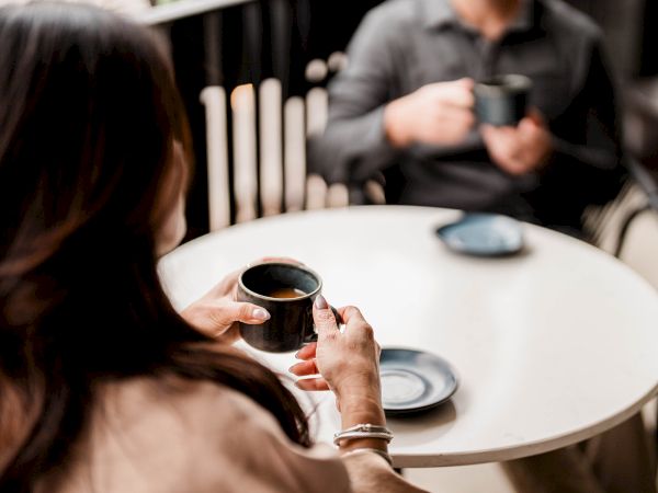A person sits at a round table holding a cup, with a saucer nearby, while another person in the background holds a mug.