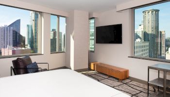 A modern hotel room with large windows showing a cityscape, featuring a bed, a wall-mounted TV, and a chair.