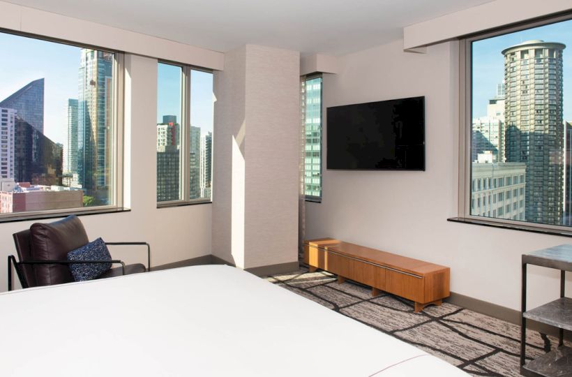 A modern hotel room with large windows showing a cityscape, featuring a bed, a wall-mounted TV, and a chair.