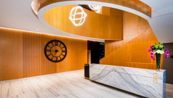 The image shows a modern lobby with a marble reception desk, wooden accents, a wall clock, and decorative lights, creating a luxurious ambiance.