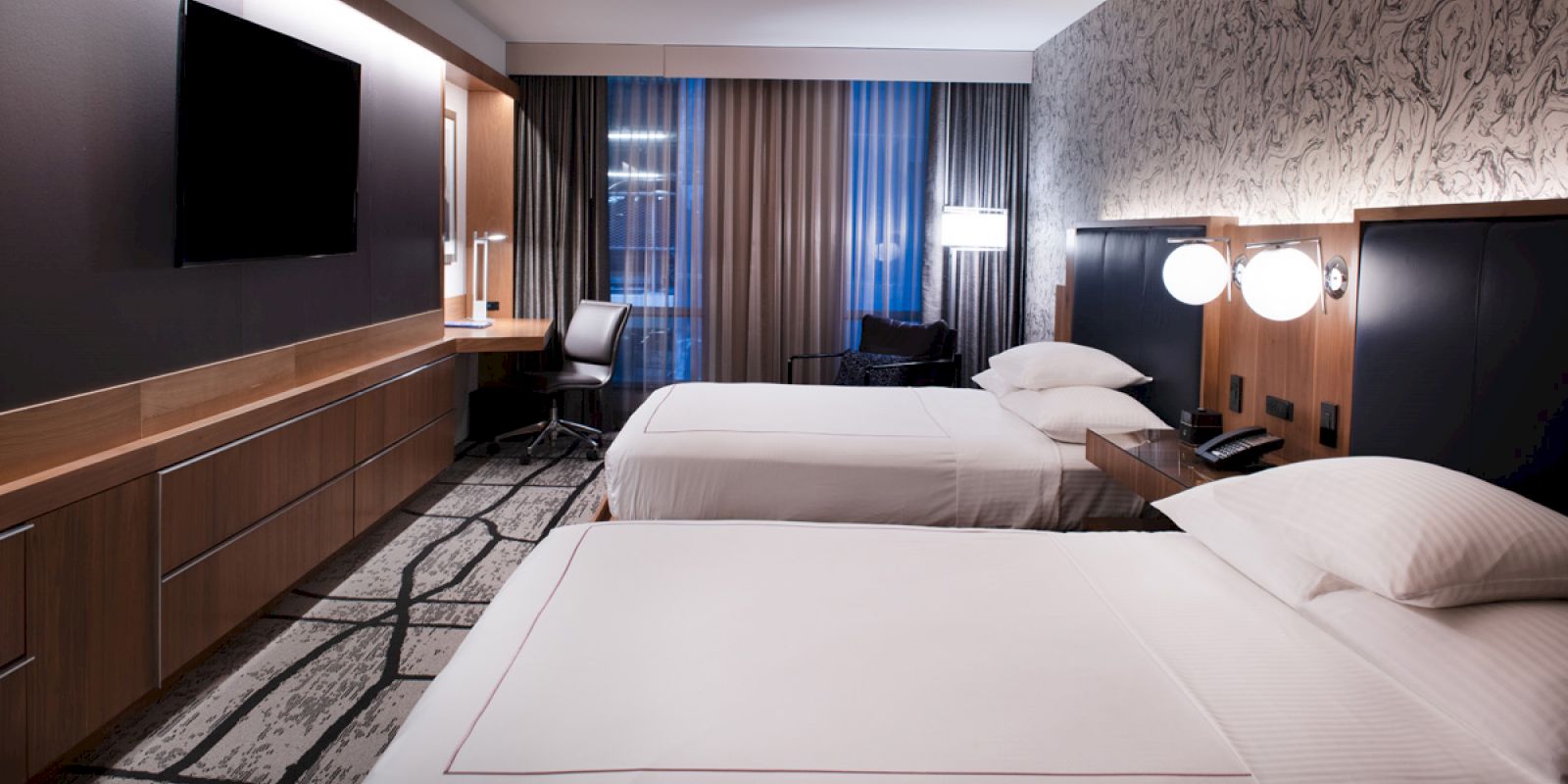 This image shows a modern hotel room with two neatly made beds, a wall-mounted TV, a desk, and decorative lighting.