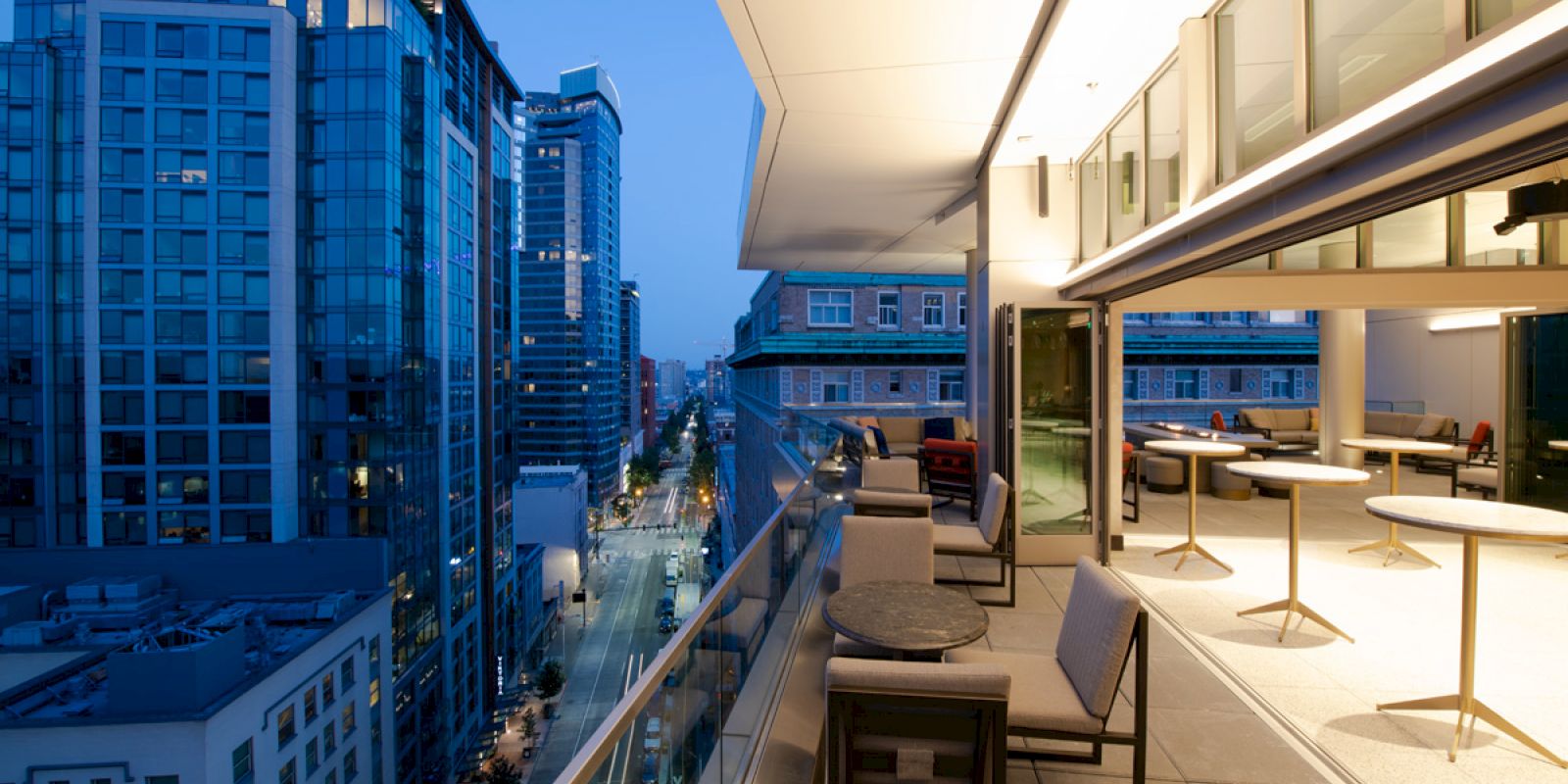 A modern rooftop terrace with seating overlooks a cityscape featuring tall buildings and a vibrant blue evening sky.