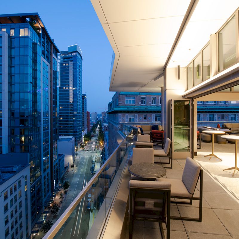 A modern, illuminated balcony with seating overlooks a cityscape during blue hour, featuring tall buildings and a serene evening atmosphere.
