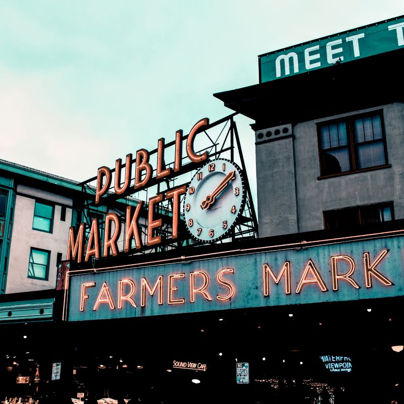 The image shows a building with a neon sign reading "Public Market Farmers Market" and a clock, under a cloudy sky.