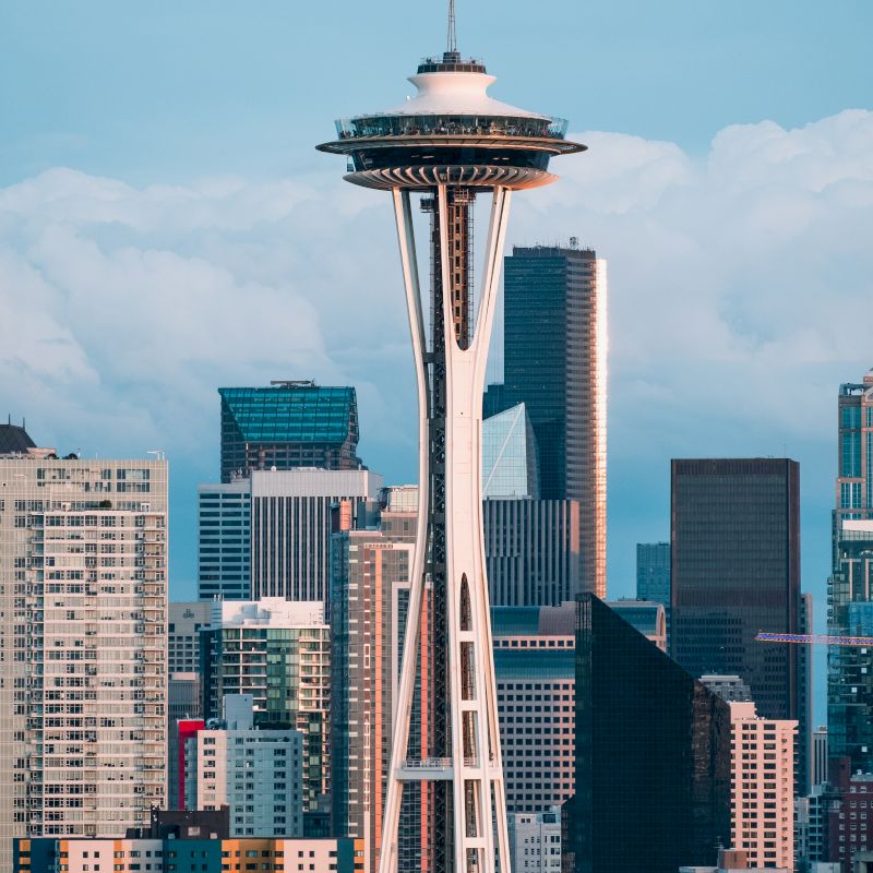 The image shows the Space Needle in Seattle, with a backdrop of modern buildings and a blue sky with clouds.