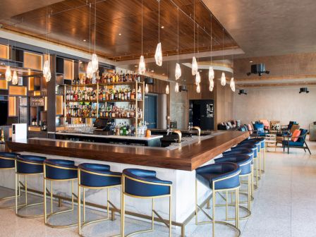 A stylish bar with modern decor, featuring high stools around a central counter, shelves stocked with bottles, and hanging lights.