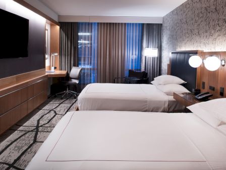 A modern hotel room features two beds, a flat-screen TV, a desk, chairs, and contemporary lighting, with gray and wood accents throughout.