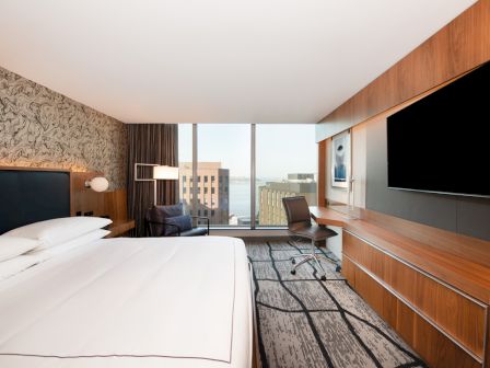 A modern hotel room with a large bed, flat-screen TV, desk, and window view of city buildings.