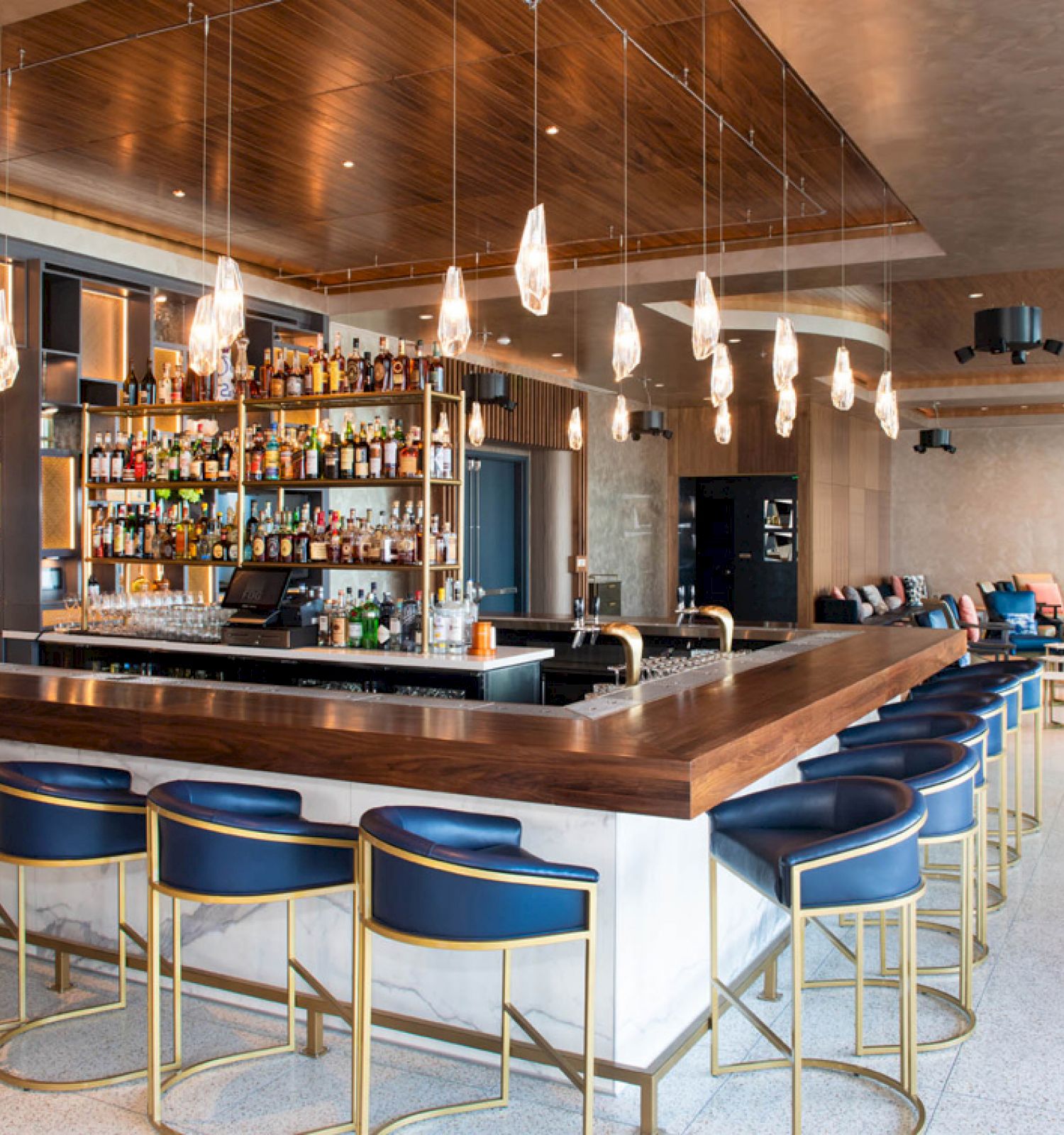 The image shows a modern bar with a wooden counter, stylish hanging lights, blue bar stools, and a well-stocked shelf in a spacious room.