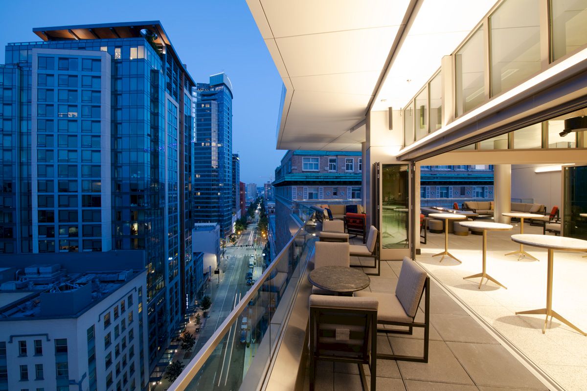A modern rooftop dining area overlooks a cityscape at dusk, featuring comfortable seating and tables amidst urban high-rises.