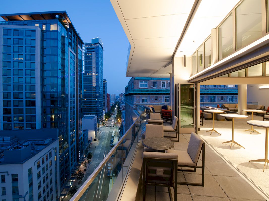 A modern rooftop dining area overlooks a cityscape at dusk, featuring comfortable seating and tables amidst urban high-rises.