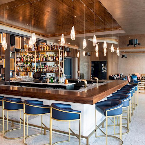 The image shows a modern bar with blue stools, hanging lights, and a well-stocked counter in an upscale lounge setting.