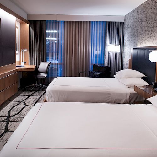 The image shows a hotel room with two beds, a TV, a desk, and ambient lighting. The decor is modern, with a patterned carpet.