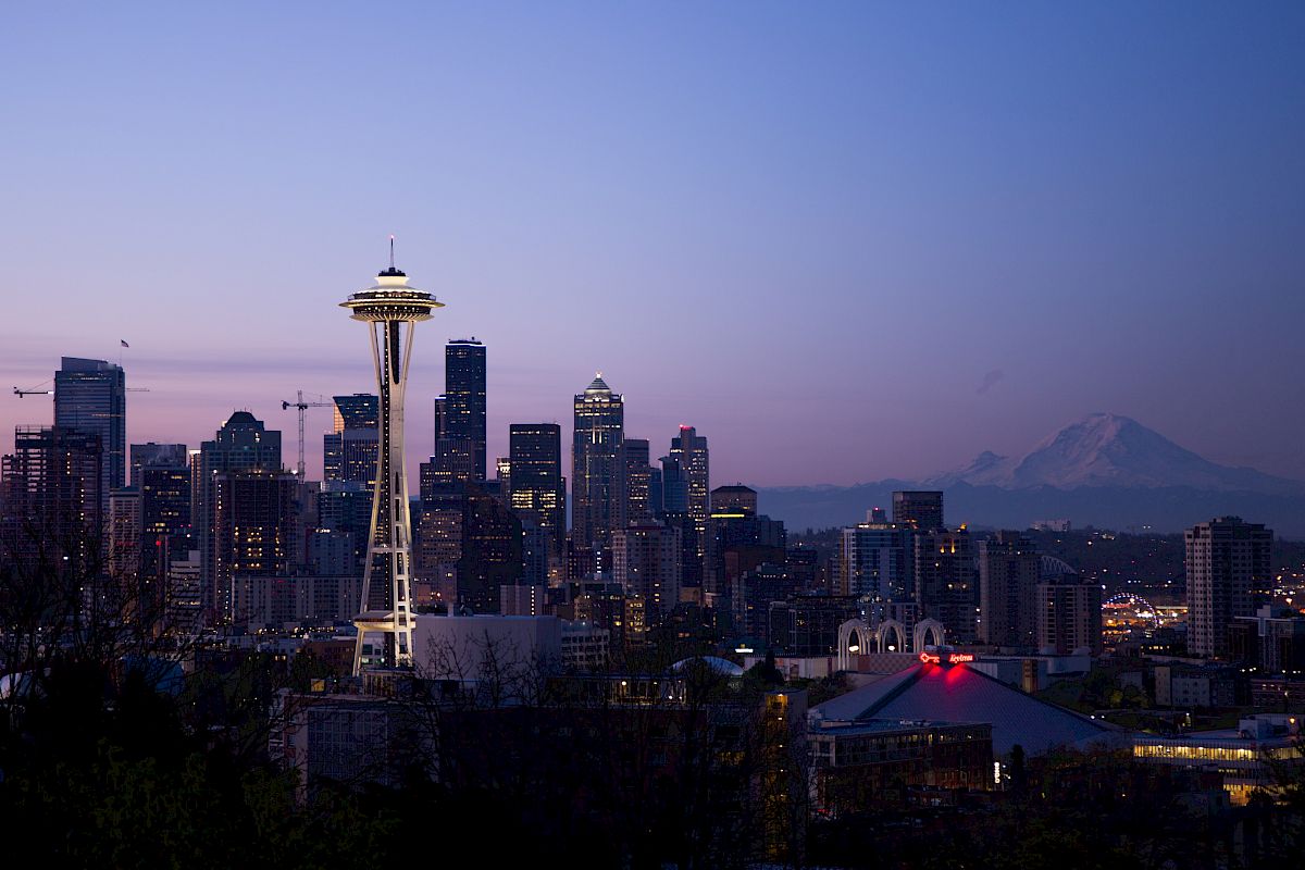 The image shows the skyline of Seattle at dusk, featuring the Space Needle and Mount Rainier in the background under a twilight sky.