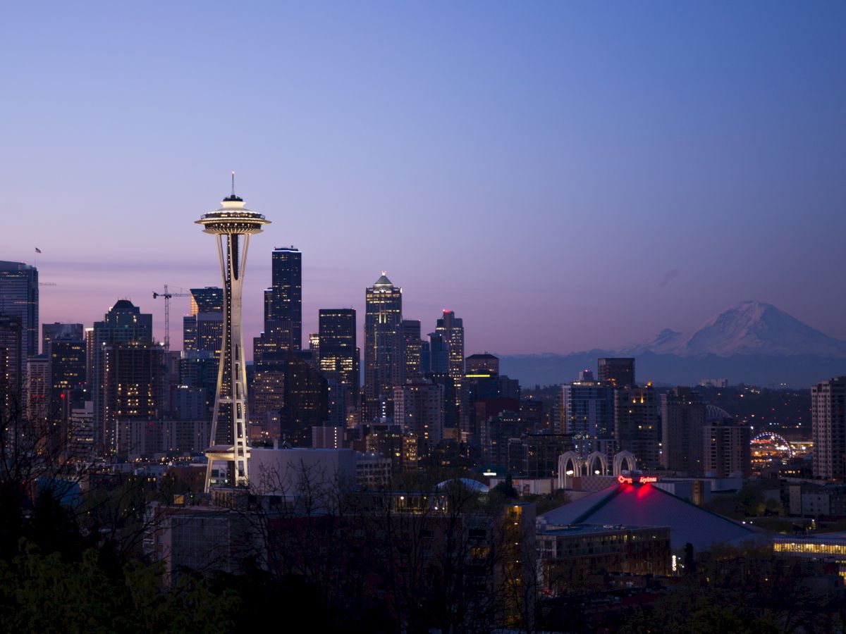 The image shows the skyline of Seattle at dusk, featuring the Space Needle and Mount Rainier in the background under a twilight sky.