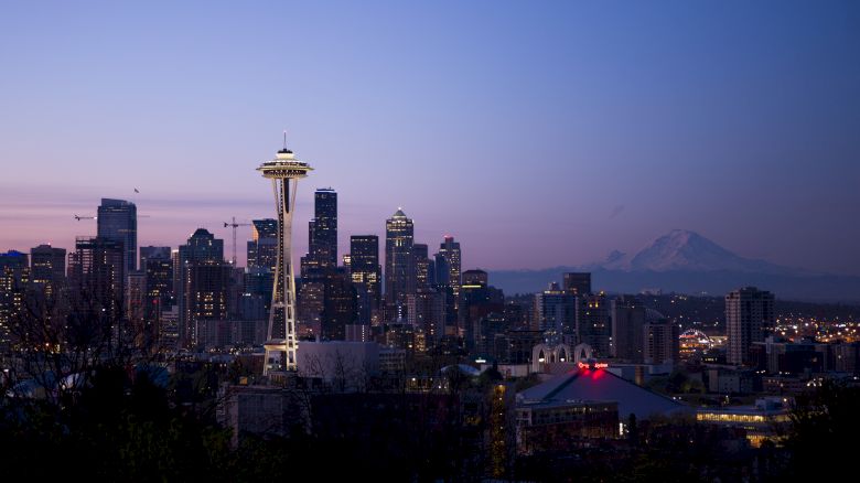 The image shows the skyline of Seattle at dusk, featuring the Space Needle and Mount Rainier in the background under a twilight sky.