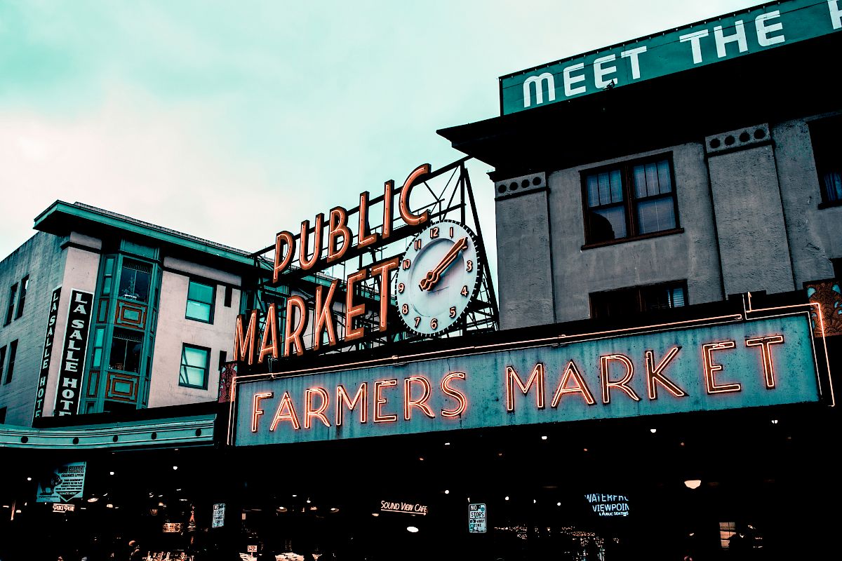 The image shows a public market with neon signs for a farmers market and a clock, set against urban buildings.
