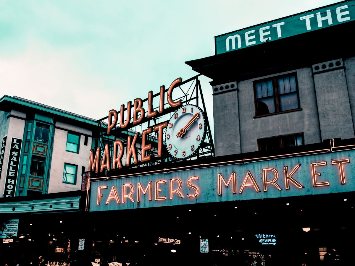 The image shows a public market with neon signs for a farmers market and a clock, set against urban buildings.