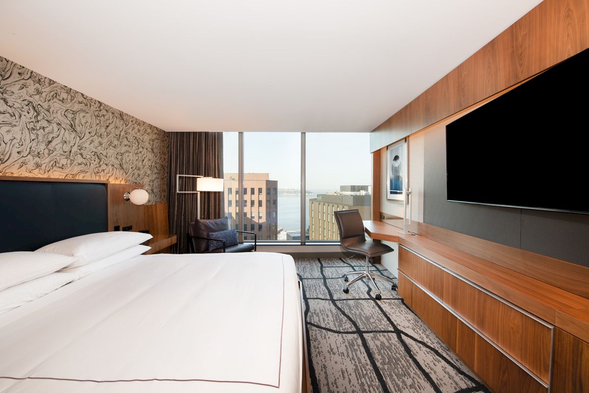 A modern hotel room with a large bed, wall-mounted TV, desk, chair, and window offering a city view, featuring sleek wooden accents.