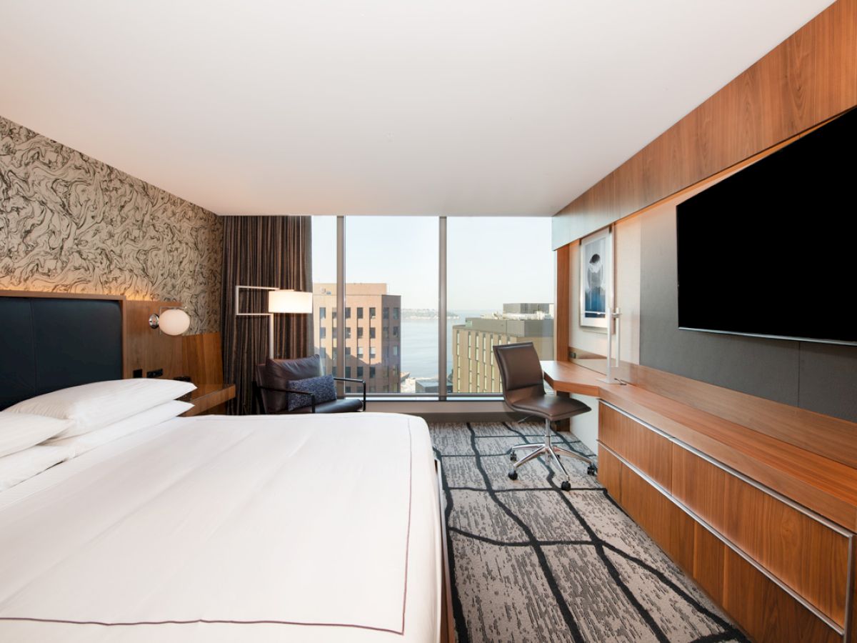 A modern hotel room with a large bed, wall-mounted TV, desk, chair, and window offering a city view, featuring sleek wooden accents.