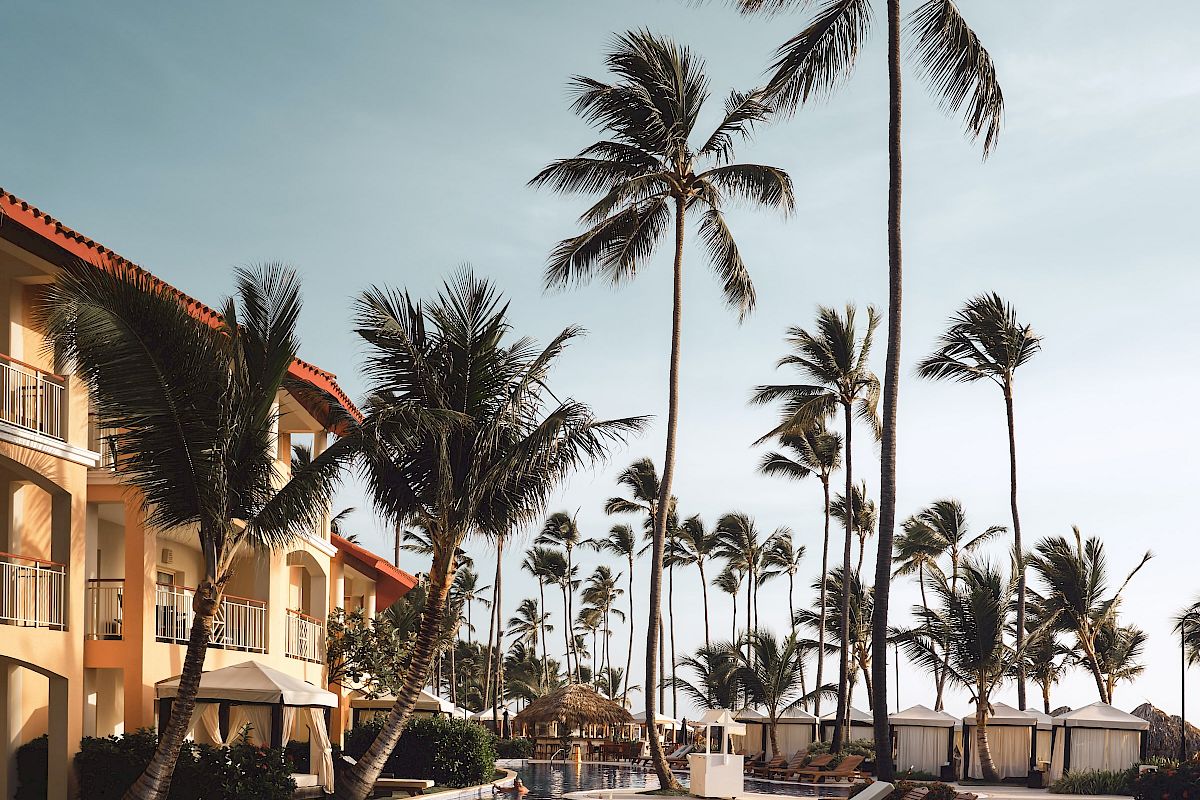 A tropical resort scene with a pool, palm trees, lounge chairs, and a building under a clear sky.
