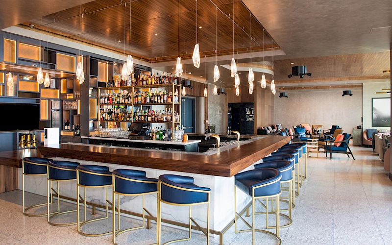 The image shows a modern bar with blue stools, pendant lighting, and a wide selection of bottles on display.
