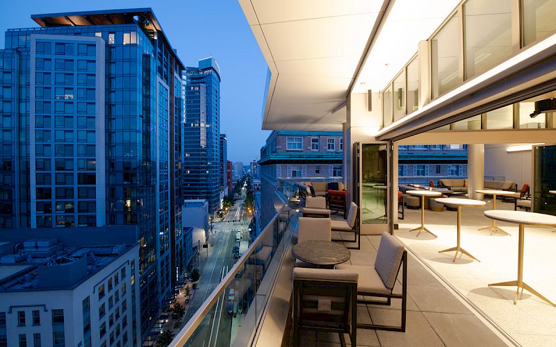 A modern balcony with seating overlooks a city street and tall buildings at twilight, creating an elegant urban atmosphere.